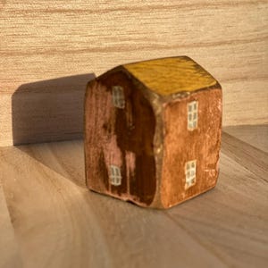 Puede incluir: Una peque&ntilde;a figura de casa de madera pintada a mano con un techo de color amarillo mostaza y paredes marrones. La estructura muestra cuatro ventanas blancas pintadas, dos en la fachada frontal y dos en el lateral, con textura de madera visible.