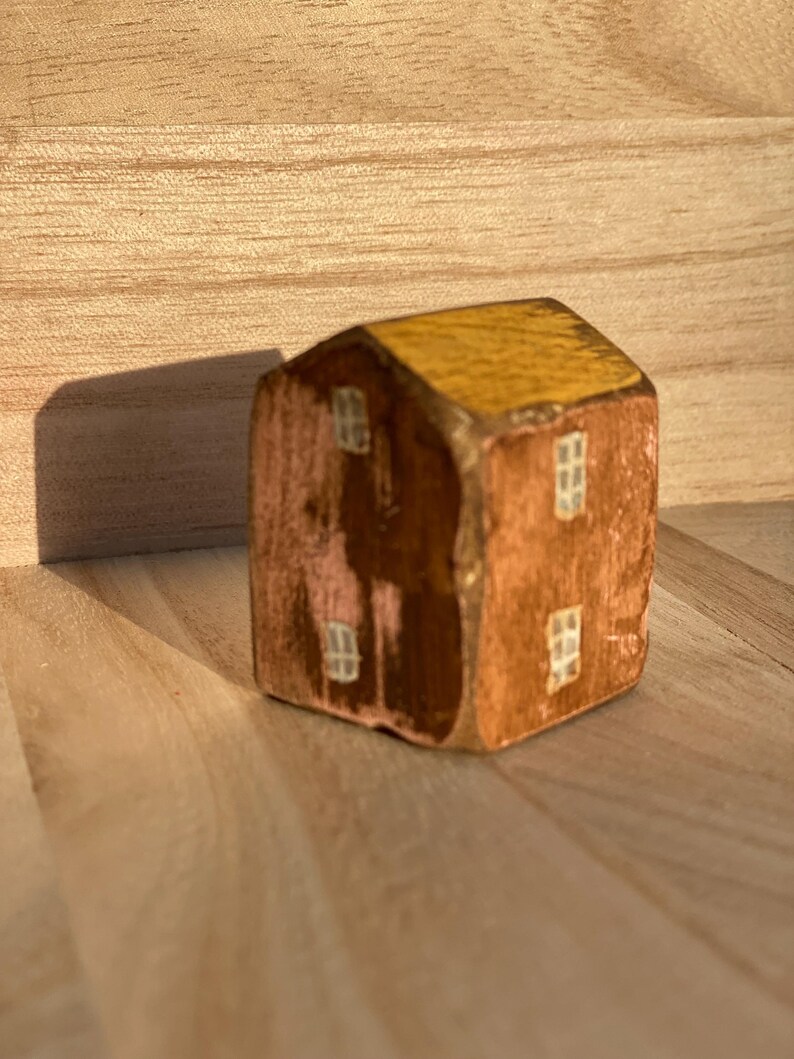 Puede incluir: Una peque&ntilde;a figura de casa de madera pintada a mano con un techo de color amarillo mostaza y paredes marrones. La estructura muestra cuatro ventanas blancas pintadas, dos en la fachada frontal y dos en el lateral, con textura de madera visible.