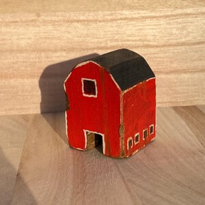 Puede incluir: Una peque&ntilde;a figura de granero de madera pintada en rojo brillante con un techo negro. La estructura presenta una ventana cuadrada con borde blanco en el frente, una abertura rectangular en la base y cuatro peque&ntilde;as ventanas rectangulares con borde blanco.