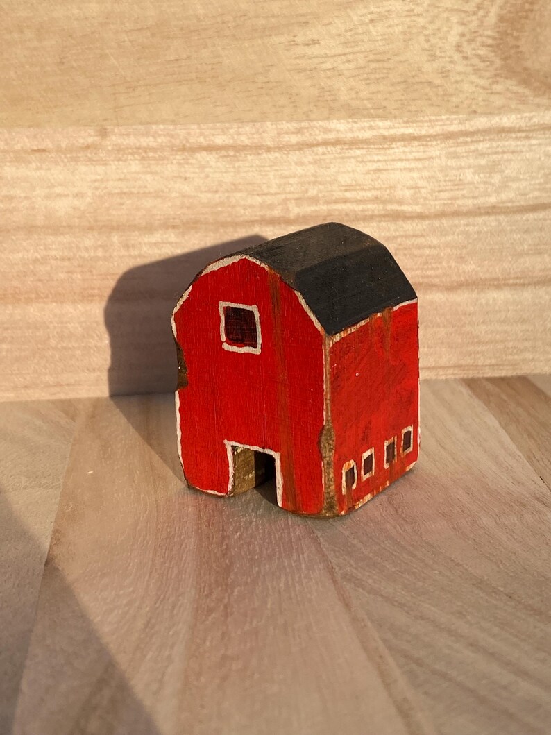 Puede incluir: Una peque&ntilde;a figura de granero de madera pintada en rojo brillante con un techo negro. La estructura presenta una ventana cuadrada con borde blanco en el frente, una abertura rectangular en la base y cuatro peque&ntilde;as ventanas rectangulares con borde blanco.