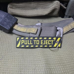 May include: A rectangular black fabric keychain featuring yellow embroidered text that reads PULL TO EJECT. The design includes yellow diagonal hazard stripes along the top and bottom borders, and a silver metal split ring is attached to one side.