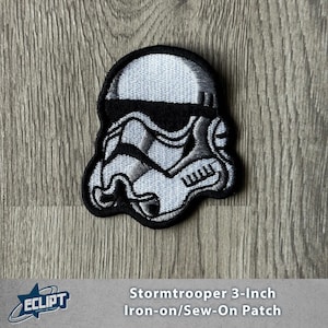 May include: A black and white Stormtrooper 7.6 cm iron-on/sew-on patch. The patch features the iconic helmet design from the Star Wars franchise, with detailed shading and a black border. The patch is on a wooden surface.