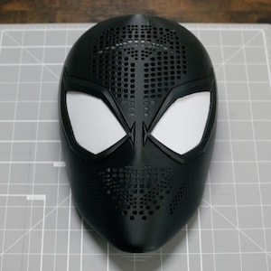May include: A matte black full-face mask featuring a perforated texture across the forehead and chin areas. The mask includes two large, angular eye openings covered with white mesh material, creating a distinct contrast against the dark surface.