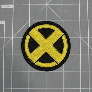 May include: A circular embroidered patch featuring a bold yellow X symbol centred on a solid black background. The design is enclosed within a thick yellow circular border, creating a high-contrast emblem with a stitched texture throughout.