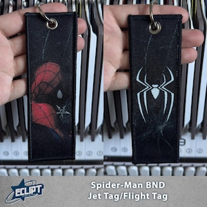 May include: A rectangular black fabric jet tag keychain featuring a red and black spider-themed mask graphic on one side and a white spider emblem on the reverse. The tag includes a silver metal grommet and a split ring for attachment.