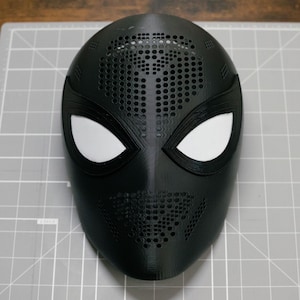May include: A black full-face mask featuring a textured surface with numerous small circular perforations across the forehead and chin areas. The mask includes two large, almond-shaped white eye lenses framed by thick black borders.