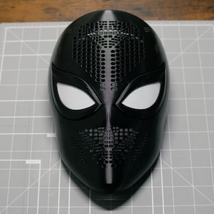 May include: A black full-face mask featuring a smooth finish with perforated sections on the forehead and nose area. The mask includes two large, almond-shaped eye openings covered with a white mesh material, creating a distinct, sleek appearance.