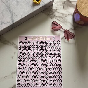 Op de afbeelding: Een roze en witte printbare kaart met zwarte hartjes en dollartekens. De kaart heeft de titel "One Dollar Saving Challenge" en heeft in totaal 120 hartjes. De tekst "Every Cent Counts" en "Saved: €120" staat onderaan de kaart.