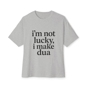 May include: A grey t-shirt featuring the black text "i'm not lucky, i make dua". The shirt has a crew neck and short sleeves. The text is centrally positioned on the front of the garment.