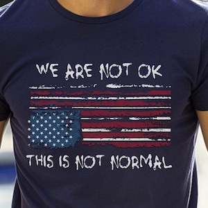 May include: Navy blue t-shirt with a distressed American flag design. The flag features red and white stripes, a blue field with white stars, and the text "WE ARE NOT OK" and "THIS IS NOT NORMAL" in white.