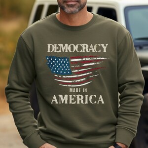 May include: A green sweatshirt with a distressed American flag design and the text "DEMOCRACY MADE IN AMERICA".