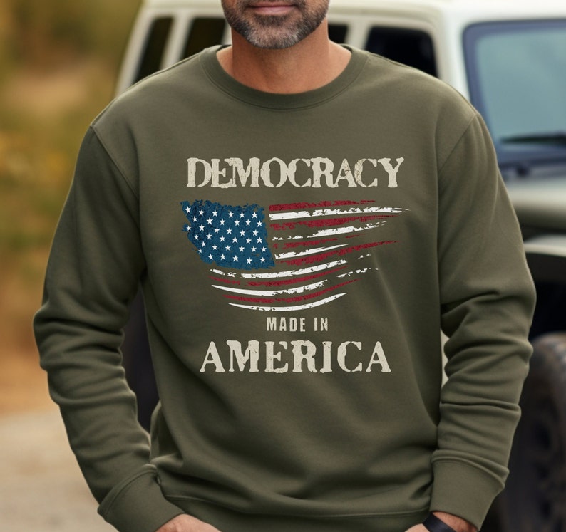 May include: A green sweatshirt with a distressed American flag design and the text "DEMOCRACY MADE IN AMERICA".
