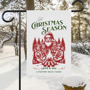 May include: A white Christmas garden flag with a black metal stand. The flag features a red and green illustration of Santa Claus with children around a campfire, with the text "The Christmas Season" and "Love & Joy".