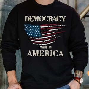 May include: Black sweatshirt with a distressed American flag design and the text "Democracy Made in America".