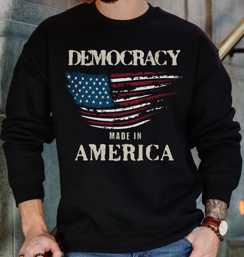 May include: Black sweatshirt with a distressed American flag design and the text "Democracy Made in America".