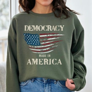 May include: A green sweatshirt with a distressed American flag design and the text "Democracy Made in America".