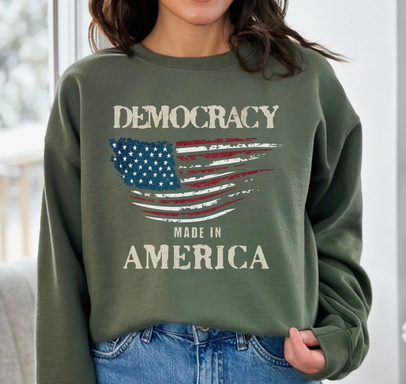 May include: A green sweatshirt with a distressed American flag design and the text "Democracy Made in America".