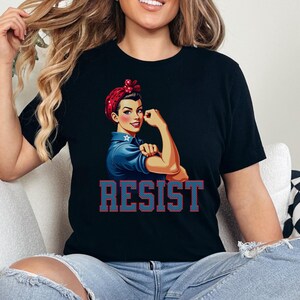 May include: Black t-shirt with a graphic of a woman with red, white, and blue hair, wearing a blue shirt and raising her fist. The text "RESIST" is printed in red and blue letters below the graphic.