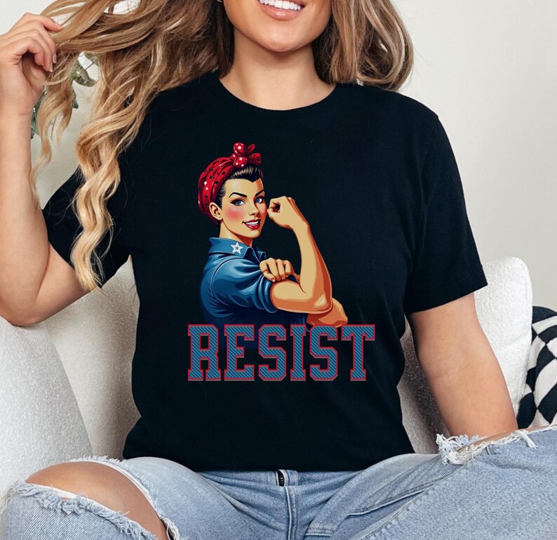 May include: Black t-shirt with a graphic of a woman with red, white, and blue hair, wearing a blue shirt and raising her fist. The text "RESIST" is printed in red and blue letters below the graphic.