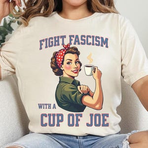 May include: Cream-coloured t-shirt with a vintage illustration of a woman wearing a red polka-dot headscarf, holding a coffee cup. The text "FIGHT FASCISM" and "WITH A CUP OF JOE" are printed in blue and red.