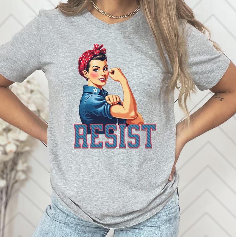 May include: Gray t-shirt with a graphic of a woman with red hair and a blue shirt flexing her bicep. The text "RESIST" is printed in red and blue letters below the graphic.