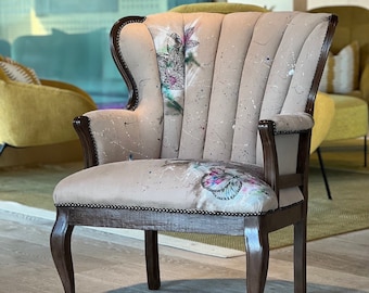 May include: A brown wingback chair with a beige fabric upholstery featuring a floral design and splattered paint. The chair has a dark wood frame and legs.