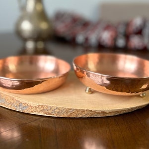May include: Two round, hammered copper bowls with a polished finish. One bowl features small, rounded metal feet on the base, while the other sits flat. Both bowls are displayed on a natural, oval-shaped wooden slice with visible bark edges.