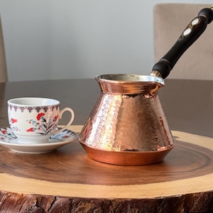 以下が含まれることがあります： 木製のトレイの上に置かれた、木製のハンドルが付いたハンマード銅製のトルコ式コーヒーポット。白い花柄のコーヒーカップとソーサーが添えられています。