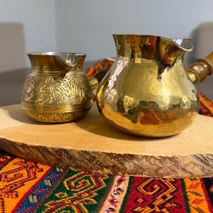 May include: Two brass coffee pots, one small with ornate floral engravings, the other larger with a wooden handle. Both pots are on a wooden serving board, with a colorful patterned textile in the foreground.