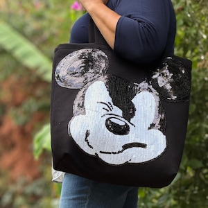 May include: Black tote bag with a large Mickey Mouse face design. The face is made with white and black sequins, with black outlining the ears and face. The bag is being carried.