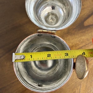 Pu&ograve; includere: Una vista dall'alto di un boccale cilindrico in metallo con coperchio incernierato. L'interno &egrave; color argento e un metro a nastro giallo &egrave; teso sull'apertura, mostrando un diametro di circa 8,9 cm. Un manico in metallo &egrave; fissato sul lato.