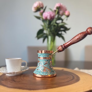 May include: A copper coffee pot with a turquoise painted body featuring intricate floral engravings and a long, dark wooden handle, accompanied by a small white ceramic cup and saucer set, showcasing a metallic finish and detailed craftsmanship.