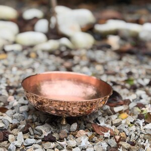 May include: A round, shallow bowl with a metallic copper finish and a hammered texture on the exterior surface. The bowl is supported by three small, rounded feet attached to the base, featuring a smooth, polished interior and a wide rim.
