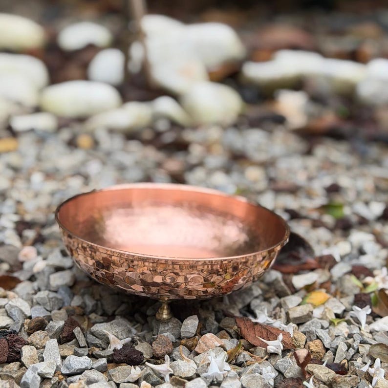 May include: A round, shallow bowl with a metallic copper finish and a hammered texture on the exterior surface. The bowl is supported by three small, rounded feet attached to the base, featuring a smooth, polished interior and a wide rim.