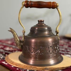 May include: A copper-toned metal kettle featuring a dark brown wooden handle, a brass-colored arched frame, and a spout with intricate embossed detailing. The body of the kettle is decorated with a wide band of engraved scrolling patterns.