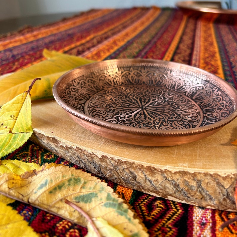 May include: A round copper bowl featuring an intricate, dark-toned engraved pattern across its interior surface. The bowl has a smooth, metallic rim and a shallow, circular shape, showcasing detailed decorative metalwork with a polished finish.