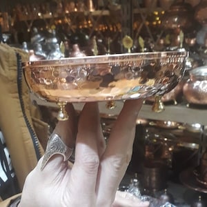 May include: A shallow, round copper bowl featuring a hammered texture on the exterior surface. The bowl is supported by three small, polished brass-colored ball feet attached to the base, providing a stable elevation for the metallic vessel.