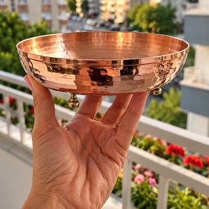 May include: A round, shallow copper bowl featuring a distinct hammered texture across its exterior surface. The bowl is supported by three small, rounded metallic feet attached to the base, providing a stable elevation for the polished copper vessel.