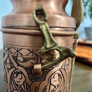 Pu&ograve; includere: Un contenitore cilindrico in rame con intricati motivi neri incisi e una finitura liscia. Una chiusura in metallo color ottone fissa il coperchio e un cucchiaio abbinato color ottone &egrave; fissato al lato del recipiente con un passante metallico.