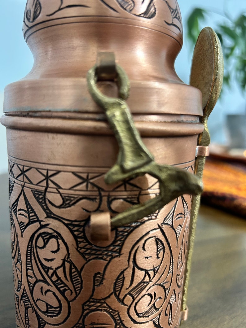 Pu&ograve; includere: Un contenitore cilindrico in rame con intricati motivi neri incisi e una finitura liscia. Una chiusura in metallo color ottone fissa il coperchio e un cucchiaio abbinato color ottone &egrave; fissato al lato del recipiente con un passante metallico.