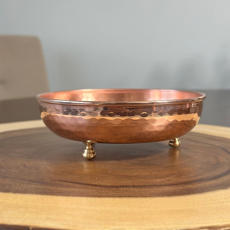 May include: A round, shallow copper bowl featuring a hammered texture on the exterior and a smooth, polished interior rim. The bowl is supported by three small, rounded brass-colored feet, providing a stable base for the metallic vessel.