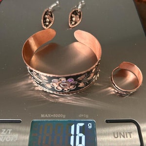 May include: A set of copper jewelry with black and white floral designs. The set includes a bracelet, a ring, and a pair of earrings. The jewelry is on a digital scale that reads 18.616 grams.