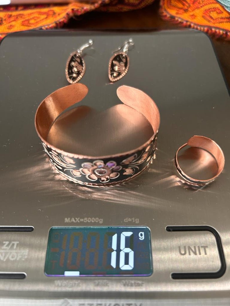 May include: A set of copper jewelry with black and white floral designs. The set includes a bracelet, a ring, and a pair of earrings. The jewelry is on a digital scale that reads 18.616 grams.