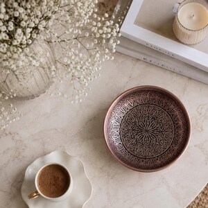 May include: A round, shallow metal bowl featuring a dark, oxidized finish with intricate, embossed floral and paisley patterns. The bowl has a smooth, metallic rim that contrasts with the detailed, textured interior design.