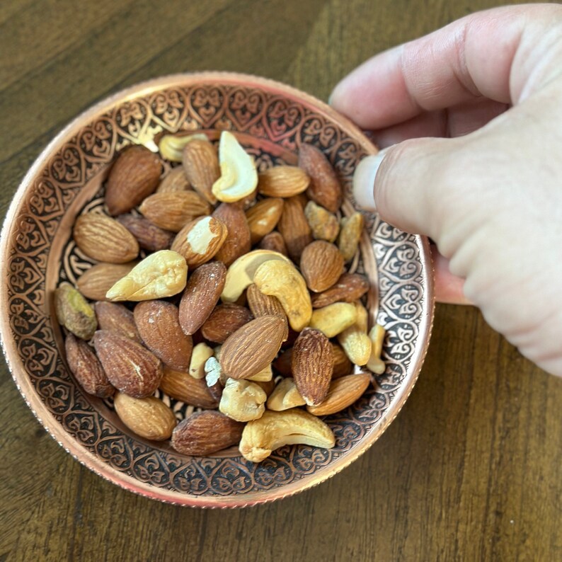 May include: A round copper-colored bowl with an intricate black etched pattern along the rim, filled with a mixture of whole brown almonds and light-colored cashew nuts. The bowl has a metallic finish and a decorative border design.