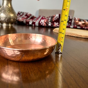 May include: A round, hammered copper bowl with a polished finish. A yellow measuring tape is positioned vertically against the side of the bowl, indicating a height of approximately 1.25 inches from the base to the rim of the metal dish.