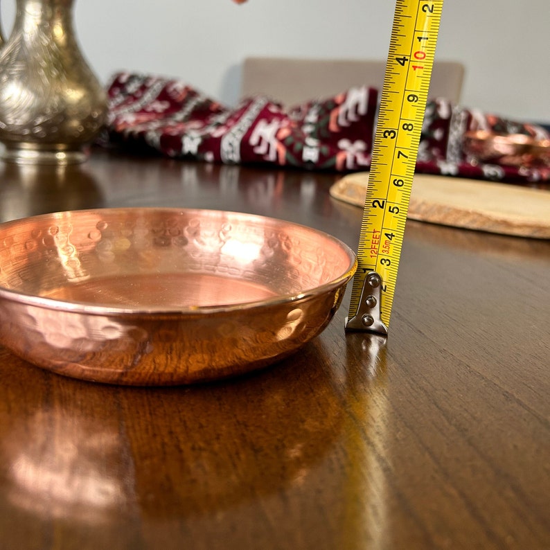 May include: A round, hammered copper bowl with a polished finish. A yellow measuring tape is positioned vertically against the side of the bowl, indicating a height of approximately 1.25 inches from the base to the rim of the metal dish.