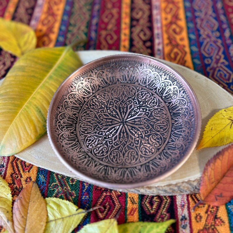 May include: A round copper bowl featuring intricate black engraved patterns across its interior surface. The design consists of symmetrical, swirling motifs radiating from the center toward the rim, creating a detailed contrast against the metallic copper.