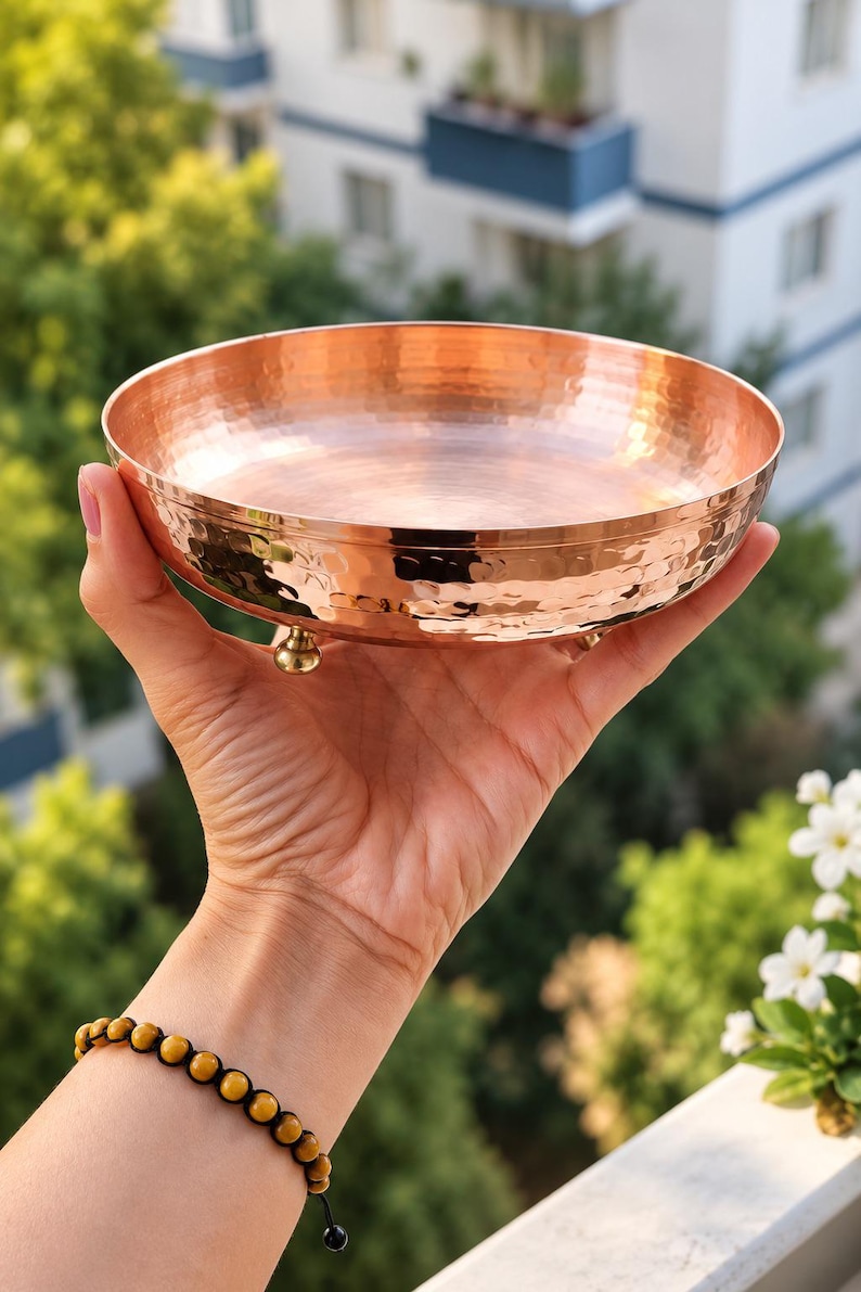 May include: A round, shallow copper bowl featuring a hammered exterior texture and a smooth, polished interior surface. The bowl is supported by three small, rounded brass-colored feet attached to the base, providing a stable elevation.