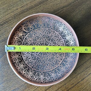 May include: A round copper-colored metal plate featuring an intricate black etched pattern of swirling lines and geometric shapes. A yellow measuring tape is placed across the center, indicating a diameter of approximately 5.5 inches.